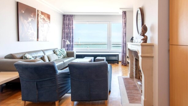 Ein modernes Wohnzimmer mit einer Couch, zwei Sesseln, einem Couchtisch und Meerblick durch große Fenster.