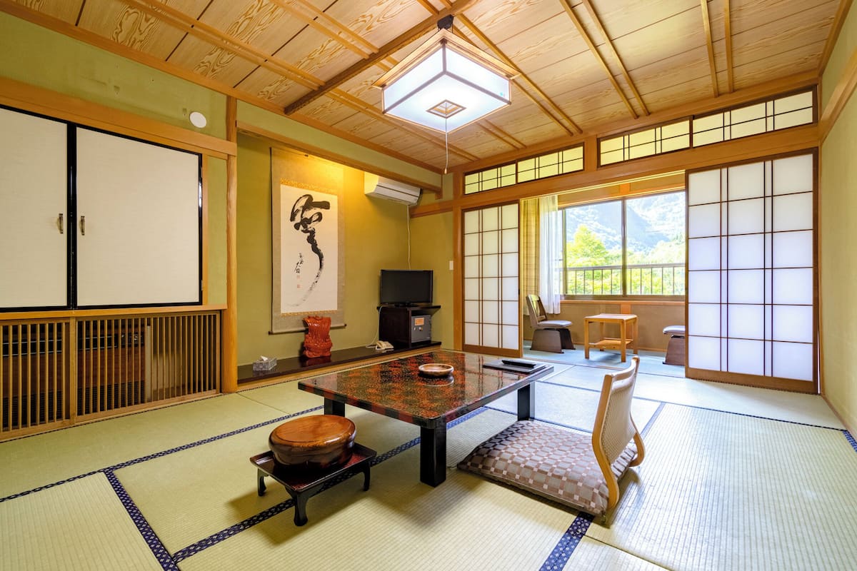 Een traditionele Japanse kamer met tatami vloerbedekking, een lage tafel, een kussenbank, schuifdeuren en uitzicht op de natuur.
