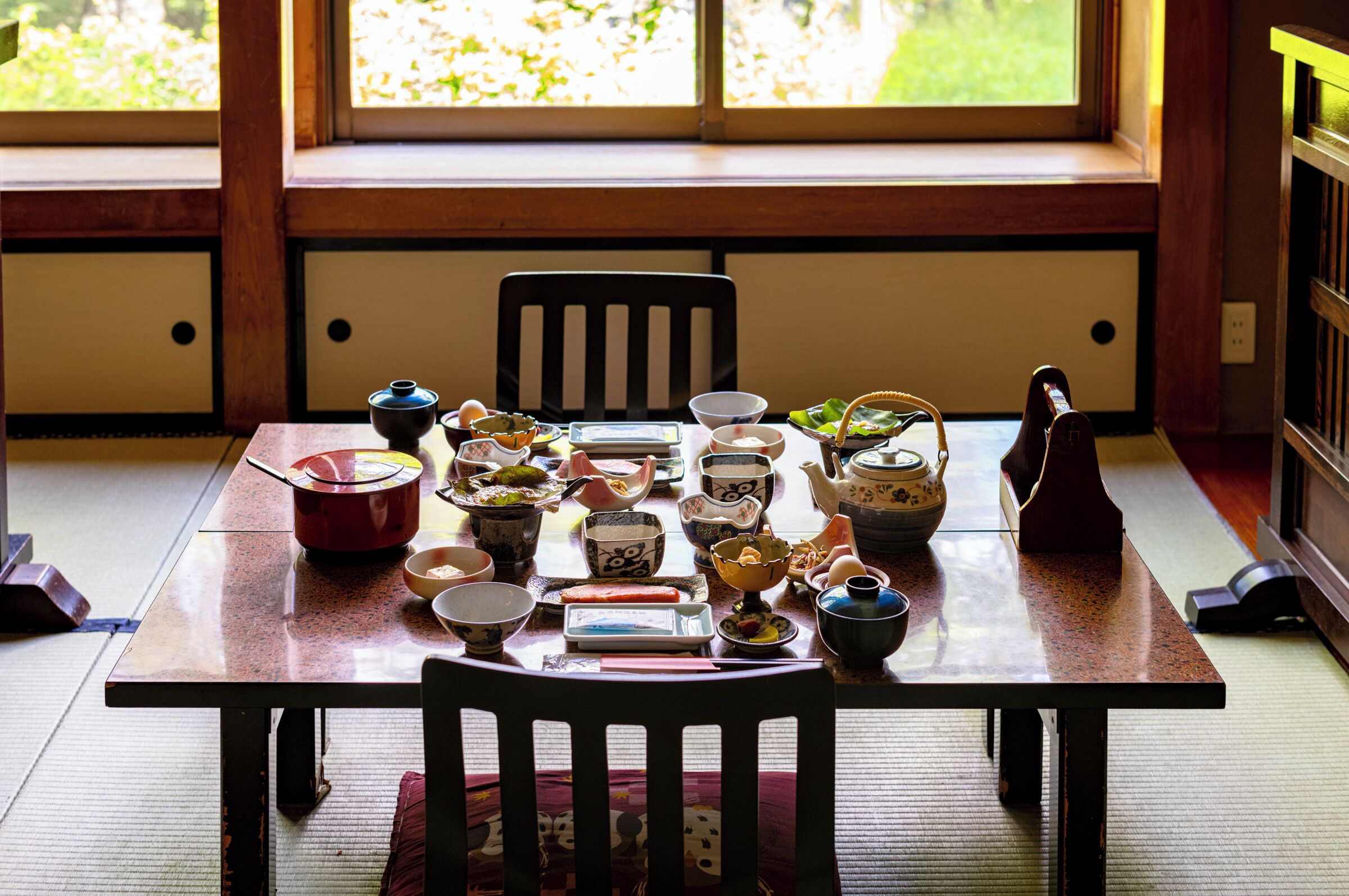 Ett traditionellt japanskt matbord med olika rätter och serveringsredskap, placerat i ett rum med tatamimattor på golvet och utsikt över grönskan genom stora fönster.