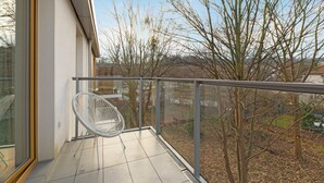 Un balcón con puerta de cristal, una silla blanca y vistas a árboles sin hojas y un edificio distante.