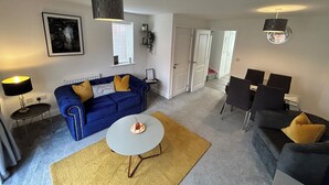 A living room with a blue sofa, a small round table, and a coffee table. There are two lamps, a clock, and a framed picture on the wall. - Radbourne House - Spacious 2 Bed, Home from Home (Derby)