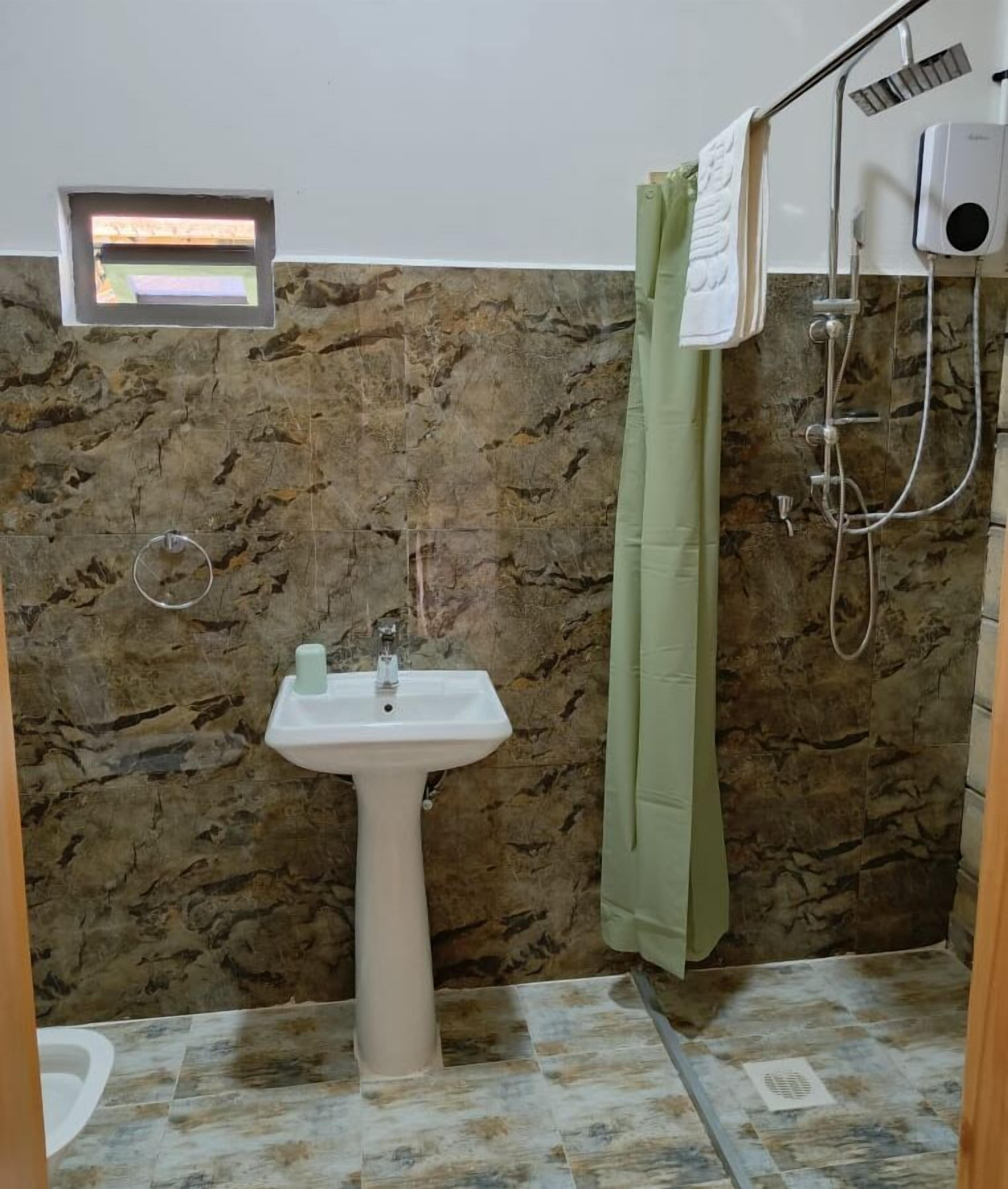 A bathroom with a pedestal sink, a shower with a green curtain, and a small window above the sink.