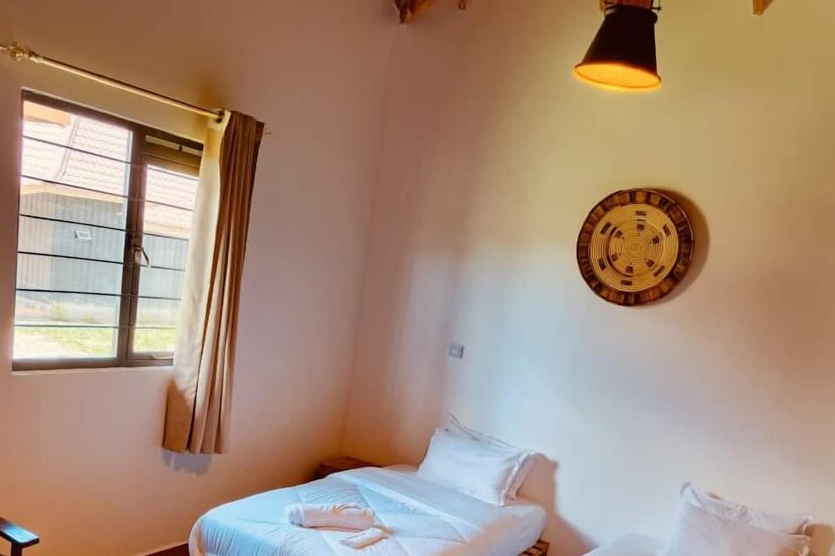 Una habitación con dos camas, piso de madera, un ventanal con cortinas y una lámpara colgante.