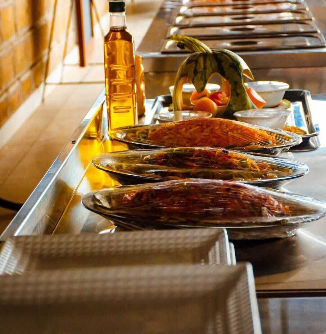 A buffet table with various dishes, a bottle of oil, and a watermelon.