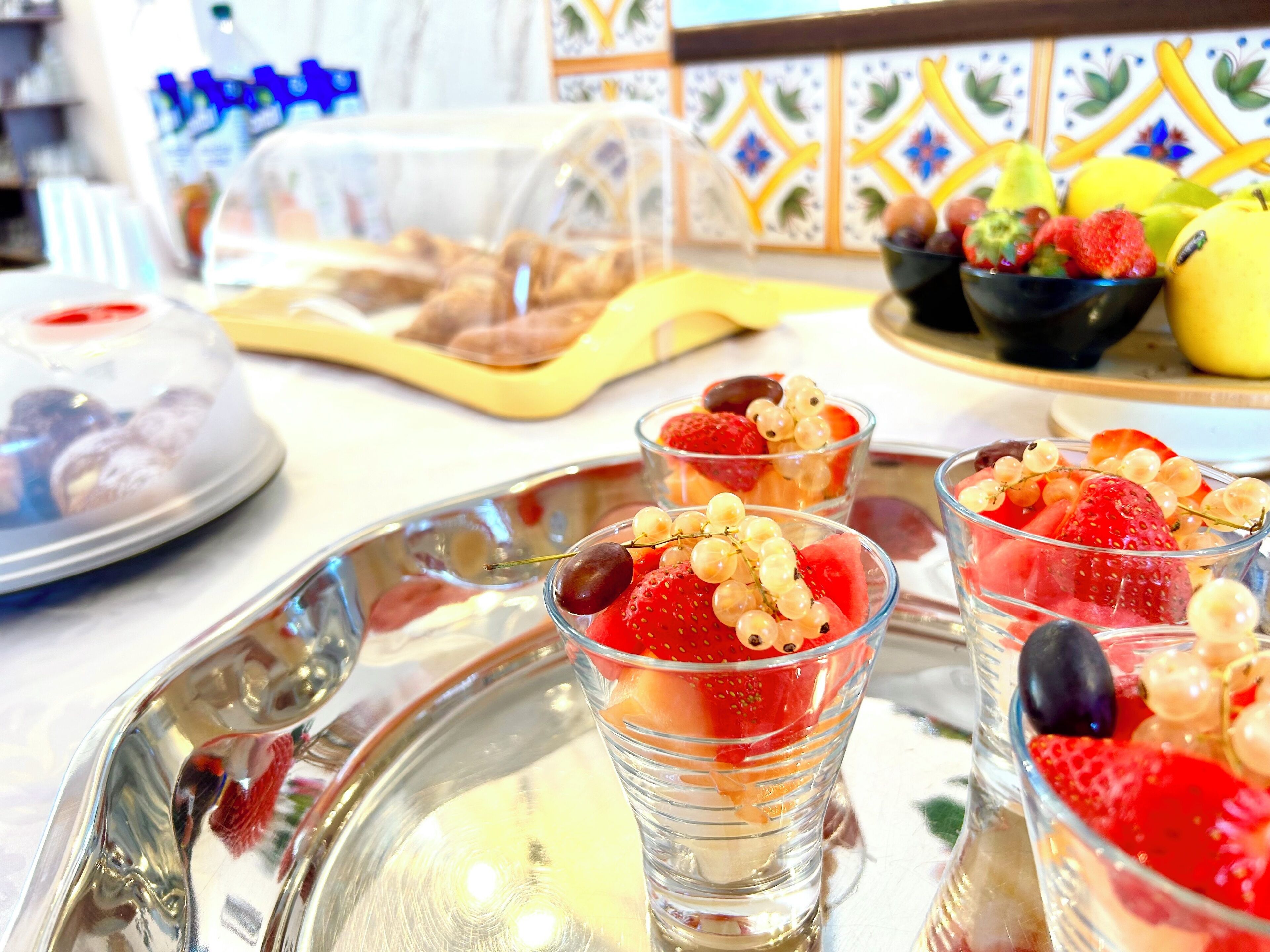 Una bandeja con tres vasos de ensalada de frutas, un bol de fruta y un recipiente con pasteles sobre una mesa.