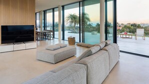 A spacious living area with a large flat-screen TV, grey sofas, a dining table, and large windows offering a view of palm trees and a pool. - Luxury villa with pool and spa - Dicasainsicilia (Riposto)