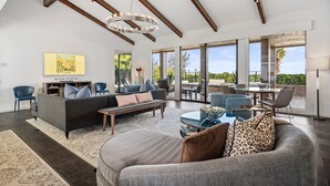 A spacious living room with a large sofa, a coffee table, and a dining area with a glass table. There is a television on the wall and a large window providing a view of the outdoors. - Ozone Pool, Spa and Built-In Bar (Palm Springs)