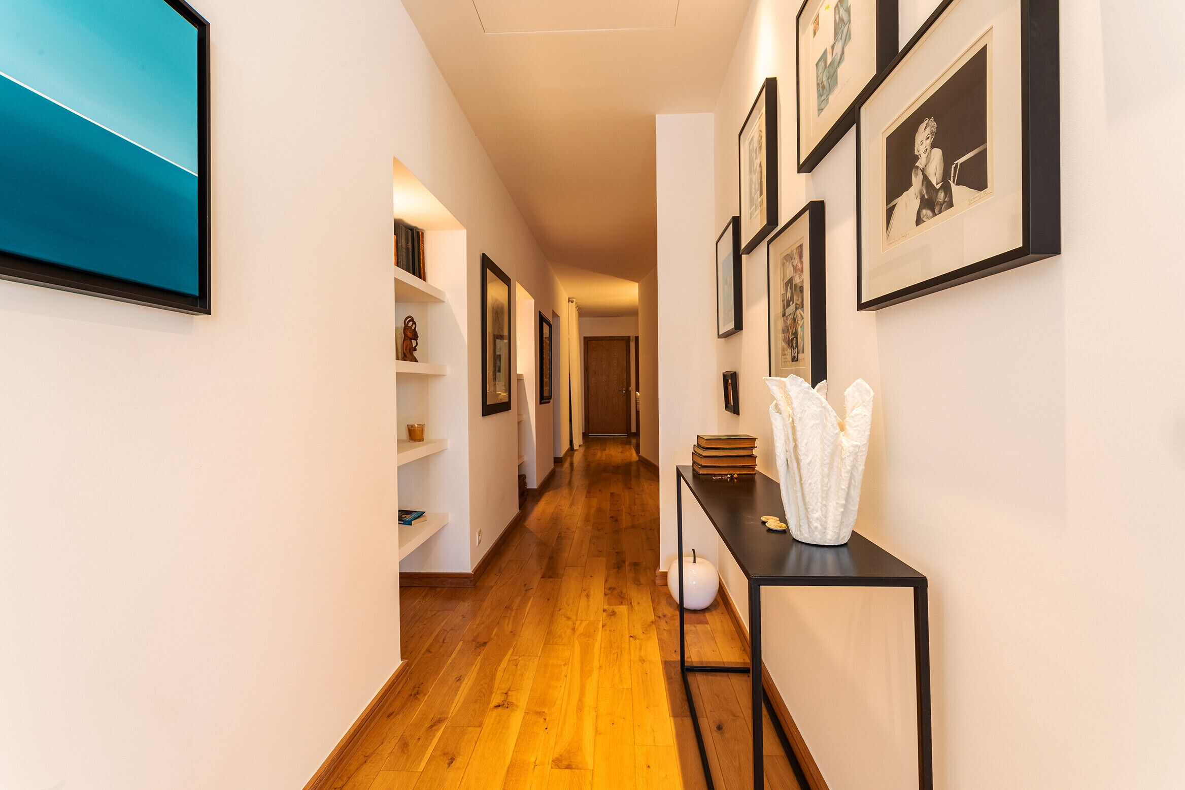 A hallway with wooden flooring, framed artwork on the walls, and a black console table with decorative items.