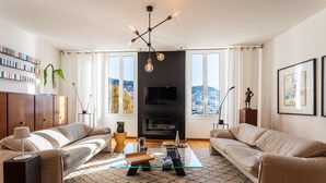 A modern living room with a sofa set, a glass coffee table, a TV, and a city view through large windows. - PORT LYMPIA AP4429 By Riviera Holiday Homes (Niza)