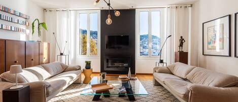 Un salon moderne comprenant un ensemble de canapés, une table basse en verre, un téléviseur et une vue sur la ville grâce à de grandes fenêtres.