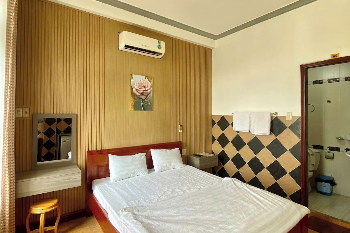 Una habitación de hotel con una cama de madera, un escritorio, un espejo y un baño visible a través de una puerta de cristal.