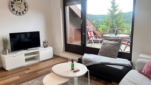 A modern living room with a television, white furniture, a round table with a bottle and glasses, and a balcony with outdoor seating. - Tannenherz vacation apartment with balcony and mountain view (Winterberg)