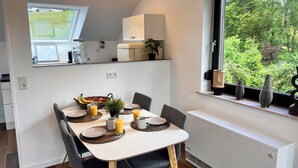 A dining area with a table set for two, a window with a view of greenery, and a white cabinet above the window. - Tannenherz vacation apartment with balcony and mountain view (Winterberg)