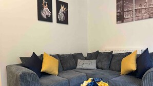 A living room with a grey striped sofa, two black and yellow cushions, a grey pillow with text, and a vase with yellow and blue flowers on a white table.