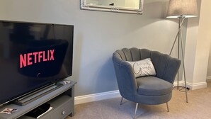 A modern living room with a large flat-screen TV displaying 'NETFLIX', a grey armchair with a cushion, and a floor lamp.