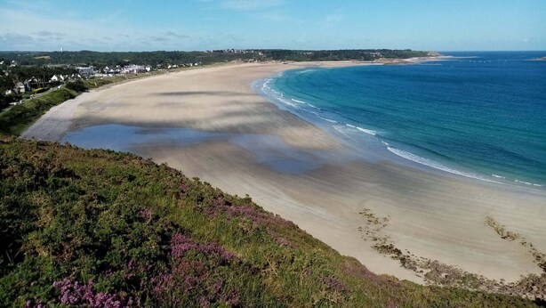 Un paysage côtier avec une plage de sable, une mer bleue et limpide, et en arrière-plan, des maisons et de la végétation.