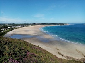A coastal landscape with a sandy beach, a clear blue sea, and a backdrop of houses and greenery.