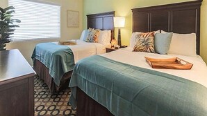 Habitación de hotel con dos camas, cada una con cabecero de madera, mesita de noche y lámpara. Hay una ventana con persianas, una planta y una alfombra estampada.