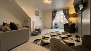 Un salon moderne avec un canapé, un fauteuil et une table basse. Un téléviseur est fixé au mur et il y a une fenêtre avec des rideaux.
