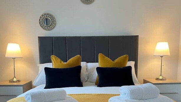 Un lit bien fait, recouvert d’une couverture jaune, avec deux oreillers et deux serviettes pliées. Deux lampes de chevet sont placées de chaque côté du lit.