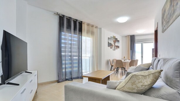 Un saló modern amb un sofà, una televisió, una taula de menjador amb cadires i una finestra gran amb cortines.