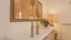 Ein Badezimmer mit weißer Ablagefläche, einem Spiegel mit Zierrahmen, einer Vase mit violetten Blumen und einer Lampe.