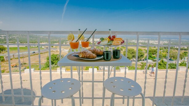 Un balcón con una mesa preparada para el desayuno, que incluye croissants, fruta y bebidas, con vistas a un paisaje pintoresco.