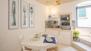 Un coin repas avec une table et des chaises blanches, une cuisine avec un mur en pierre et une fenêtre avec des volets bleus.