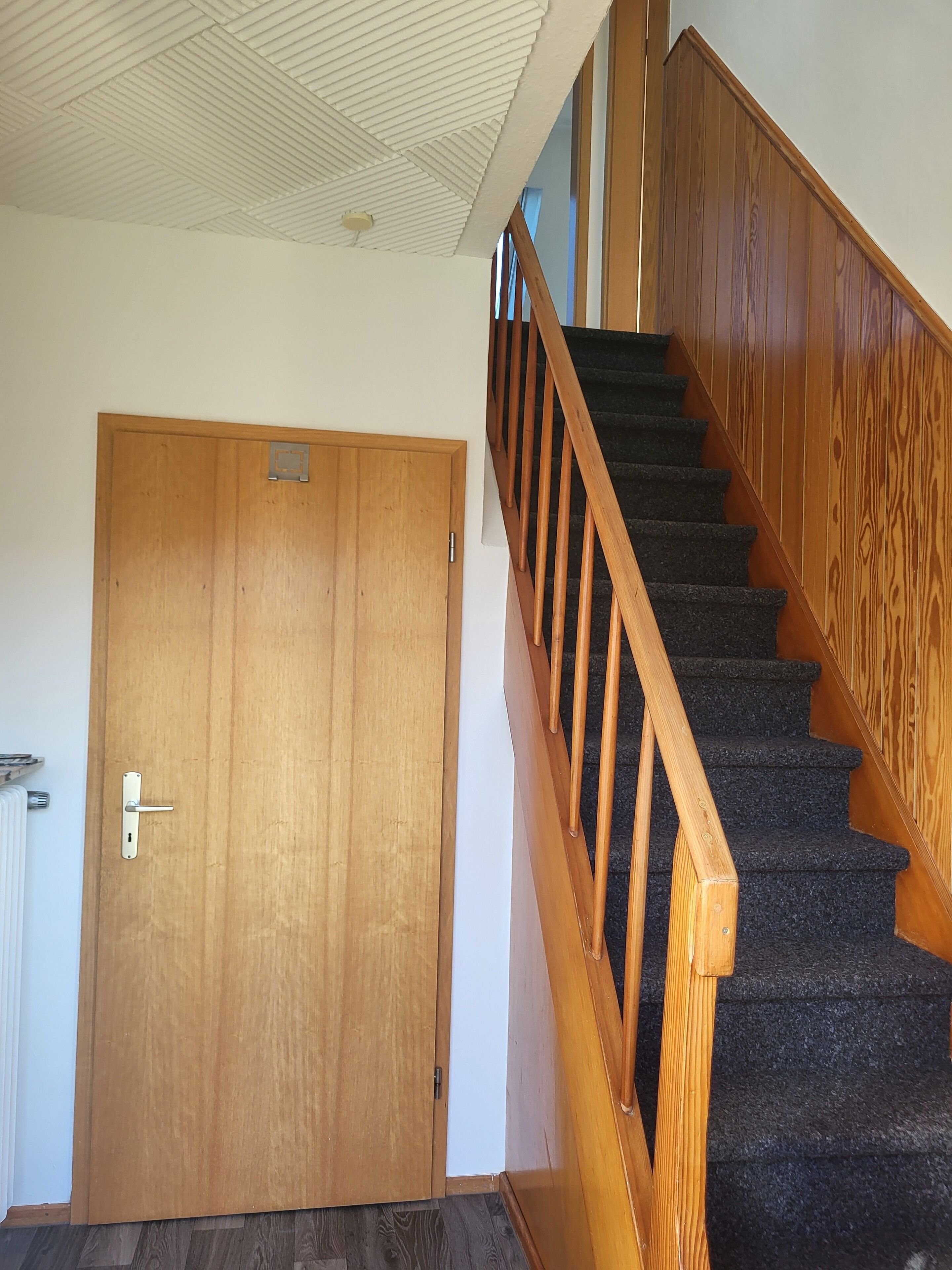 Une porte en bois avec une poignée en métal et un trou de serrure, un escalier en bois avec une rampe, et un plafond blanc orné d’un motif quadrillé.