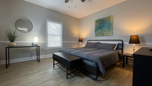 Ein Schlafzimmer mit Bett, Nachttischen, einer Bank, einem Spiegel, einem Fenster mit Jalousien und einem Bild an der Wand.