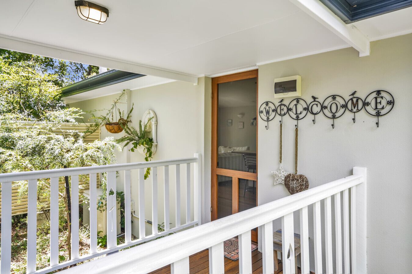 A porch with a white railing, potted plants, and a wall-mounted welcome sign.