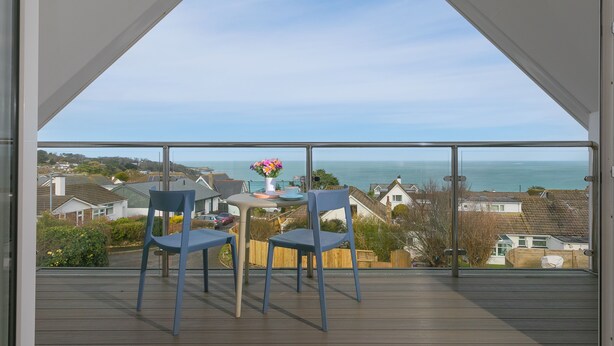 A balcony with two chairs and a small table with a vase of flowers, overlooking a residential area and the sea. - Solstice, Carbis Bay – Sleeps 8 – Sea Views – Luxury Hot Tub – On-Site Parking (Carbis Bay)