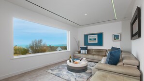 A modern living room with a large window, a round coffee table, a beige sofa, and a blue accent pillow. - Solstice, Carbis Bay – Sleeps 8 – Sea Views – Luxury Hot Tub – On-Site Parking (Carbis Bay)