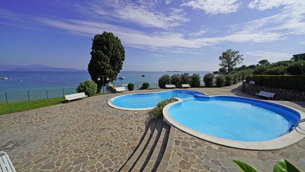 A swimming pool area with two pools, stone flooring, and a view of the sea. - Specchio sul Lago in Manerba del Garda (Manerba del Garda)