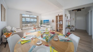 A modern living room with a dining area set for a meal, featuring a large window with a city view, a sofa, a coffee table, and a bookshelf. - Sky Suite in the Heart of the City (Fort-de-France)