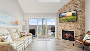 A living room with a sofa, a fireplace, a TV mounted on a stone wall, and a balcony with a view. - Out of Office - Lakeside Pool, Elevator, & Location! (Osage Beach)