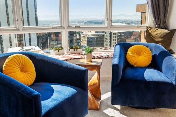 Un salon moderne avec deux fauteuils bleus, une table basse en bois et une vue sur le paysage urbain, visible à travers de grandes fenêtres.