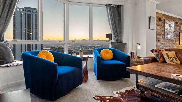 Un salon moderne avec deux fauteuils bleus, une table basse en bois et une vue sur la ville à travers de grandes fenêtres.