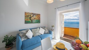 A bright living room with a blue sofa, a painting, a potted plant, and a view of the ocean through a balcony door. - Romantic cottage with outdoor heated whirlpool and breathtaking views (Praiano)