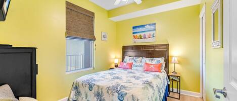 Une chambre à coucher comprenant un lit, des lampes de chevet, un ventilateur de plafond, une fenêtre avec des stores et une porte.