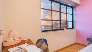 Ein Zimmer mit einem Holztisch, einem pinken Korb voller Bücher, einem Stuhl mit gemustertem Sitzkissen und einem Fenster mit Blick auf den Himmel und Palmen.