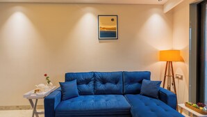Ein modernes Wohnzimmer mit einem blauen Modulsessel, einem kleinen Beistelltisch mit einer Teekanne und einer Rose sowie einer Stehlampe.