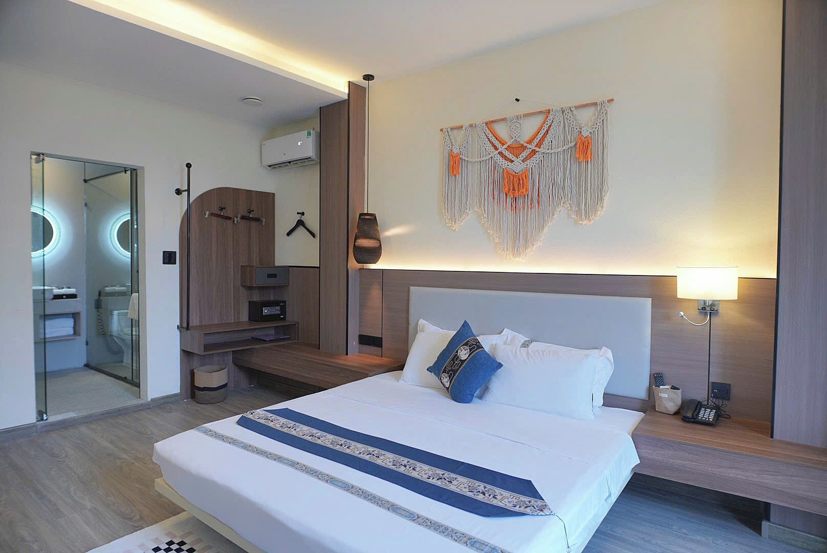 A modern hotel room with a large bed, bedside tables, a wall-mounted macrame decoration, a hanging lamp, and a mirror.