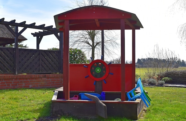 A children's play structure with a red roof and a steering wheel design, surrounded by a brick wall and a grassy area.