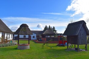 Un terrain de jeux avec des balançoires et un toboggan, entouré de constructions à toits de chaume et d’un bâtiment blanc avec une porte rouge.