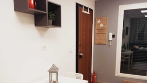 A small, well-lit room with a white table, two chairs, a small shelf with a red object, a door, a wall-mounted phone, and a window with blinds.