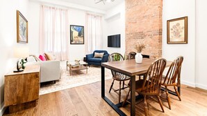 A modern living room with a dining area, featuring a brick wall, wooden furniture, and artwork on the walls.