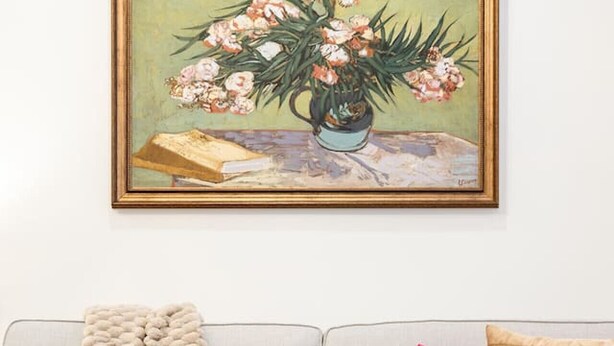 A living room with a grey sofa, a beige throw blanket, a pink pillow, a glass coffee table with a bowl and books, and a framed painting above.