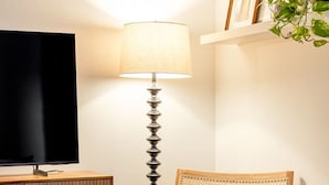 Een woonkamer met een televisie, een lamp, een rieten stoel, een tafeltje en een boekenplank.