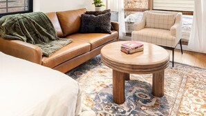 A modern living room with a brown leather sofa, a round coffee table, and a patterned rug. There is a large window with curtains, a brick wall, and a ceiling fan.